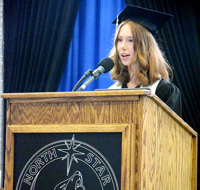 Young graduate in cap and gown speaks at a wooden podium with a mic, before blue and black curtains and a North Star emblem.