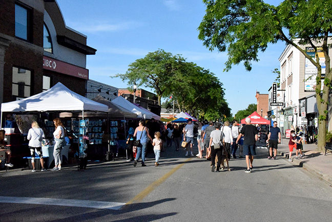 People stroll past white vendor tents at a sunny street fair lined with trees and shops, including a Vertical Centre sign.