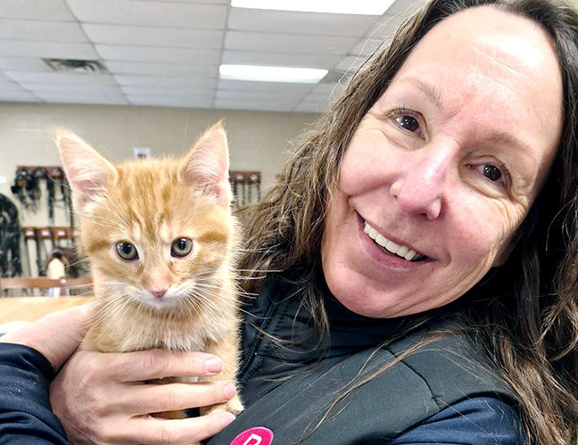 Smiling woman holds an orange kitten in a room with a tiled ceiling. The kitten looks curious. Neutral-colored background.