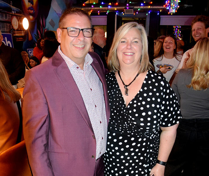 Smiling man in a purple suit and woman in a black polka-dot dress pose at a lively indoor party with colorful string lights.