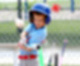 Young boy in a blue helmet swings a bat at a baseball on a field, with chain-link fence and bats in the background.