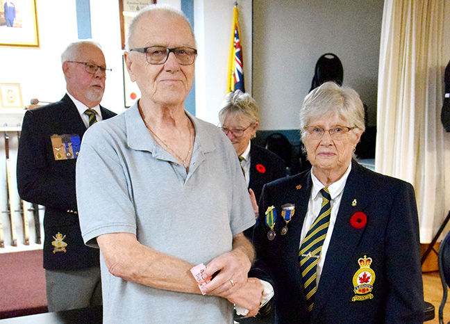 Elderly man in a polo shirt shakes hands with a medal-wearing woman at an indoor veterans ceremony, others and flags behind.