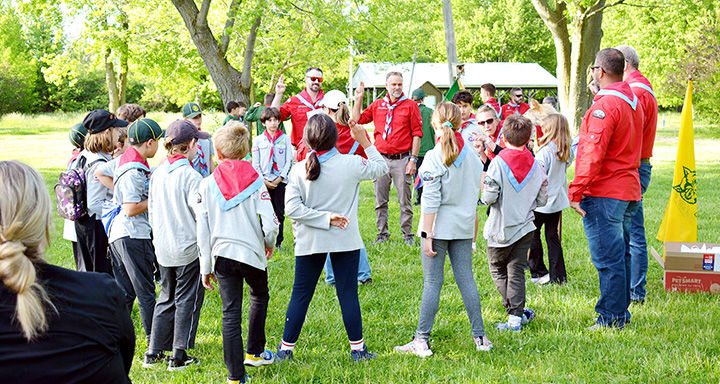 Scouts and leaders in red and gray uniforms gather in a sunny park, listening to a speaker near a yellow flag.
