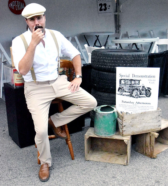 Man in white cap and shirt sits on a stool beside vintage tires and a poster reading Special Demonstration at an outdoor display.