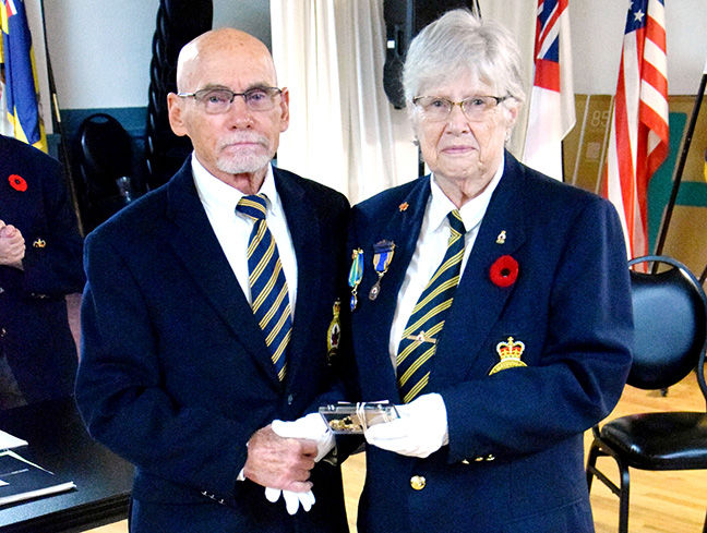 Elderly man and woman in navy uniforms with medals and white gloves hold a plaque at a formal ceremony, with flags behind them.