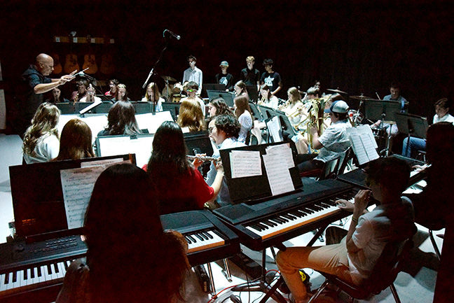 An orchestra rehearses in a dimly lit room with keyboards and sheet music. A conductor guides musicians playing various instruments.