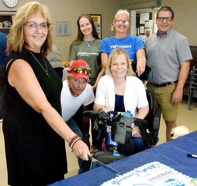 Group of smiling adults gathered around a cake at a celebration, with shirts reading Habitat for Humanity and YN'Y Variety.
