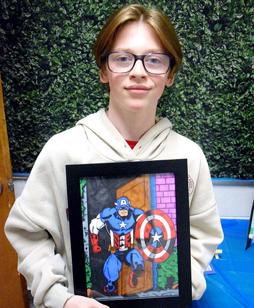 Boy in glasses and hoodie holds Captain America drawing. Green leafy background, blue table. Proud expression.