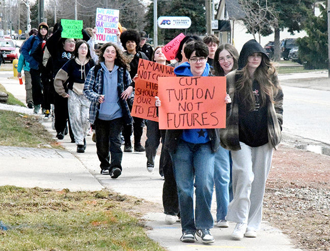 A group of students march on a sidewalk holding protest signs reading "Cut tuition not futures" and more. The mood is determined.