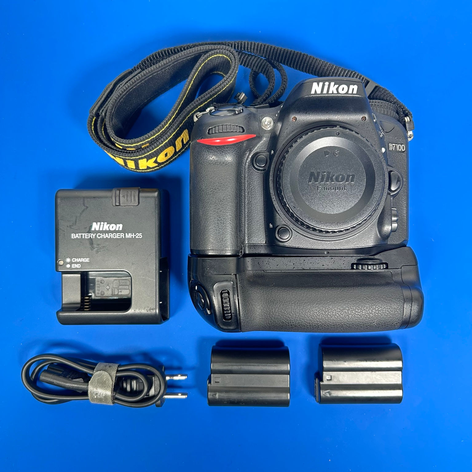 Nikon D7100 DSLR camera body kit with battery grip, two batteries, MH-25 charger, power cord, strap, and body cap