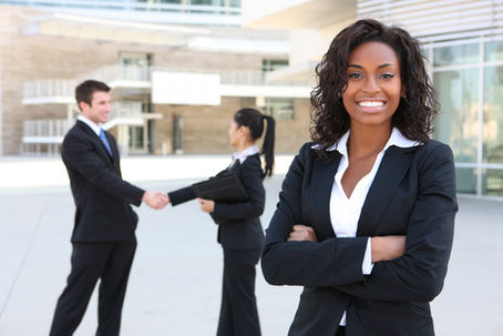 Business woman in suite crosses arms and looks at camera. Behind her, a man and women in suits shake hands