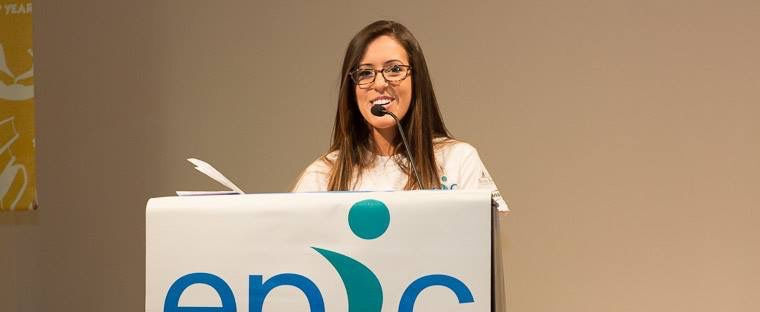 A woman wearing glasses is standing at a podium with a microphone