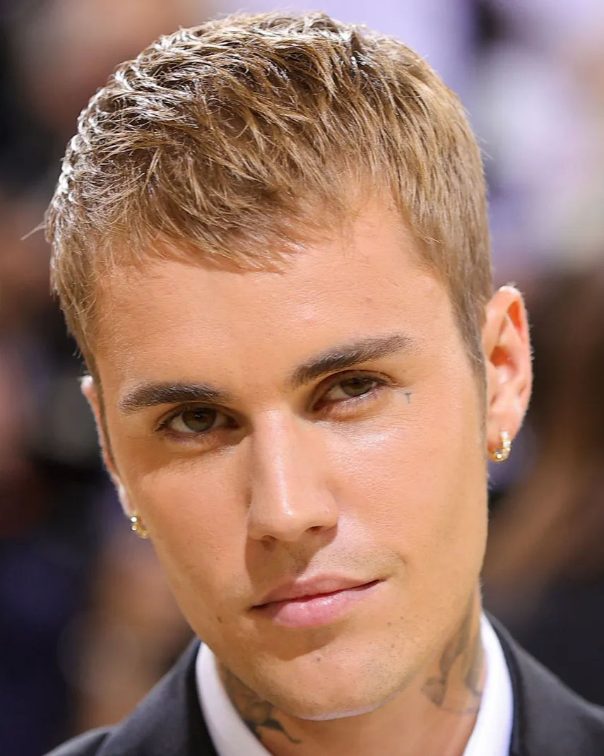 Justin Bieber wearing a dark suit and white shirt at a red carpet event, looking directly at the camera with short bleached-blonde hair, visible neck tattoos, and stud earrings.