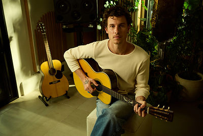 Shawn Mendes holding a Martin acoustic guitar while seated indoors, with a second guitar on a stand beside him and greenery in the background.