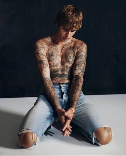 Justin Bieber shirtless, showing off his chest and arm tattoos, seated on the floor