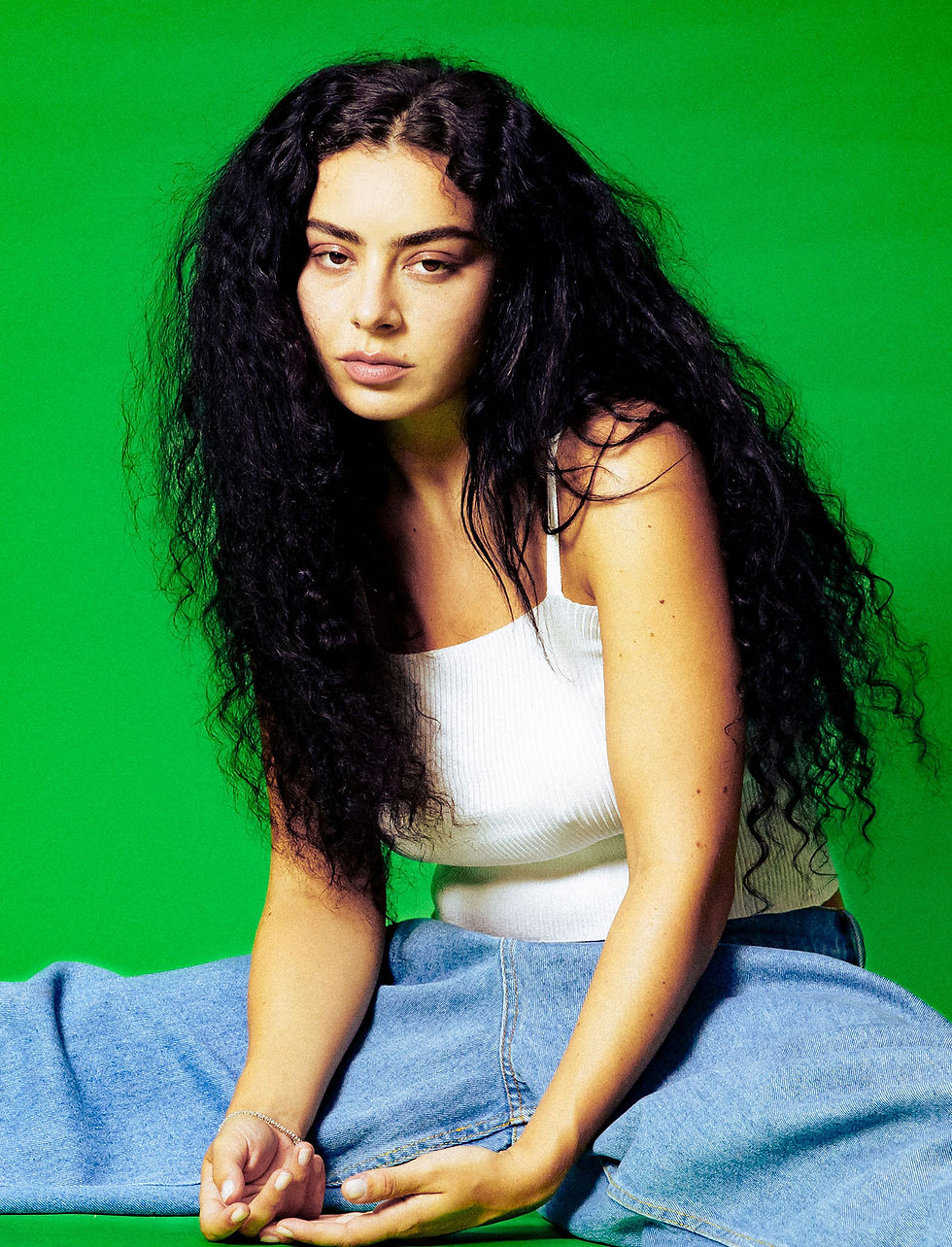 Charli XCX poses against a bright green background in a white ribbed tank top and denim, with long curly dark hair, for a 2024 press photo shoot.
