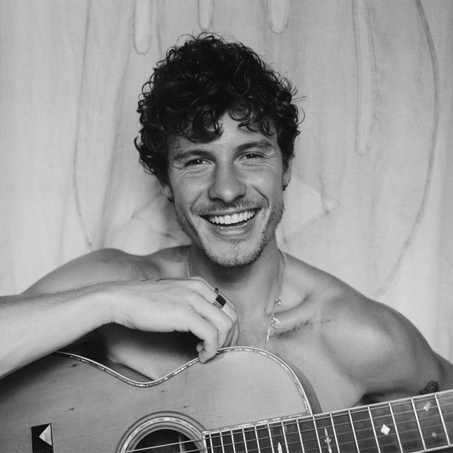 Black-and-white portrait of Shawn Mendes shirtless, leaning on an acoustic guitar, laughing with curly hair tousled and a necklace visible against a soft draped backdrop.