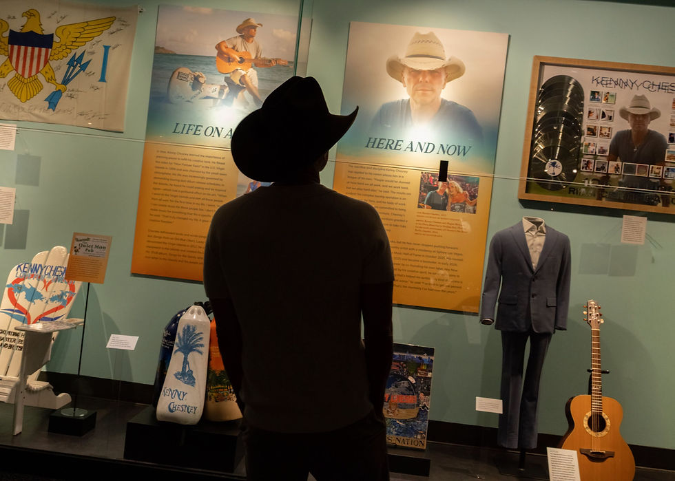 Kenny Chesney in a black cowboy hat, seen in silhouette from behind, views a museum display titled "Here and Now" featuring a large portrait of Kenny Chesney in a straw hat. The surrounding case holds memorabilia including a signed U.S. Virgin Islands flag, a beach chair, a framed collage of photos and vinyl records, a tailored suit on a mannequin, and an acoustic guitar.