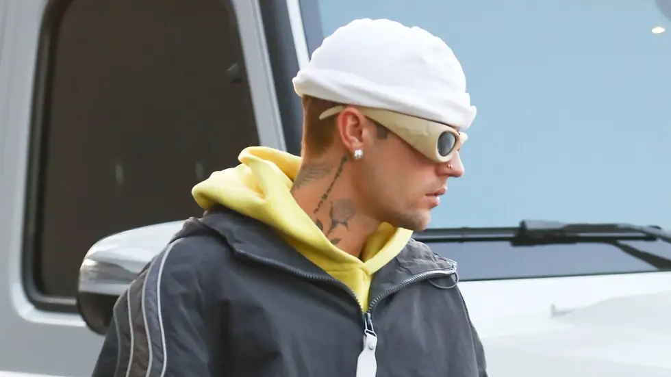 Justin Bieber wears a white beanie, tinted wraparound sunglasses, and a yellow hoodie under a gray zip-up jacket while walking outside near a parked vehicle.