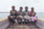Four women sit on a wooden boat holding seaweed, smiling. They're surrounded by water, wearing colorful, patterned skirts. Cloudy sky.