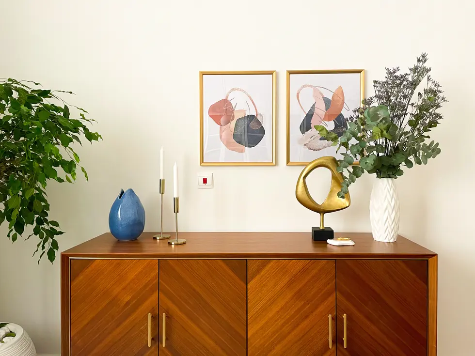 Stylish home office design by Miriam Roberts from the Roberts Renovations portfolio, featuring a mid-century modern wooden sideboard with chevron patterns and brass handles. The sideboard is elegantly decorated with abstract art prints in gold frames, a gold sculptural ornament, a white textured vase with eucalyptus branches, and a blue ceramic vase. Brass candle holders with white taper candles add a touch of sophistication, while a potted tree in a white planter introduces natural elements to the space.