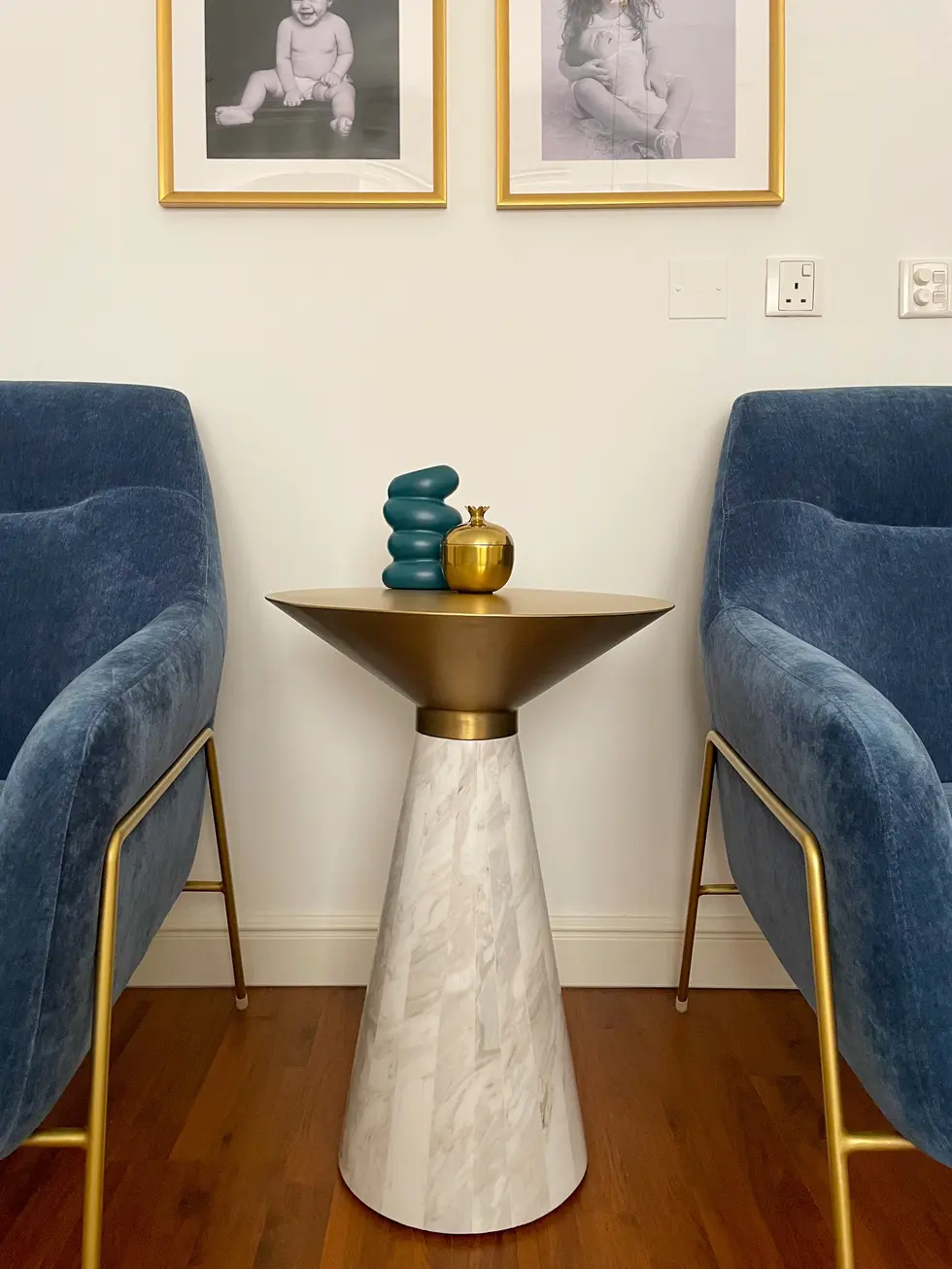 Elegant home office design by Miriam Roberts from the Roberts Renovations portfolio, featuring a sophisticated seating area with two blue velvet chairs accented by brass legs. A marble and brass cone-shaped side table sits between the chairs, adorned with a sculptural teal vase and a small gold ornament. The backdrop showcases two black-and-white family portraits in gold frames, adding a personal and warm touch to the space. The rich wooden flooring complements the luxurious yet inviting atmosphere.