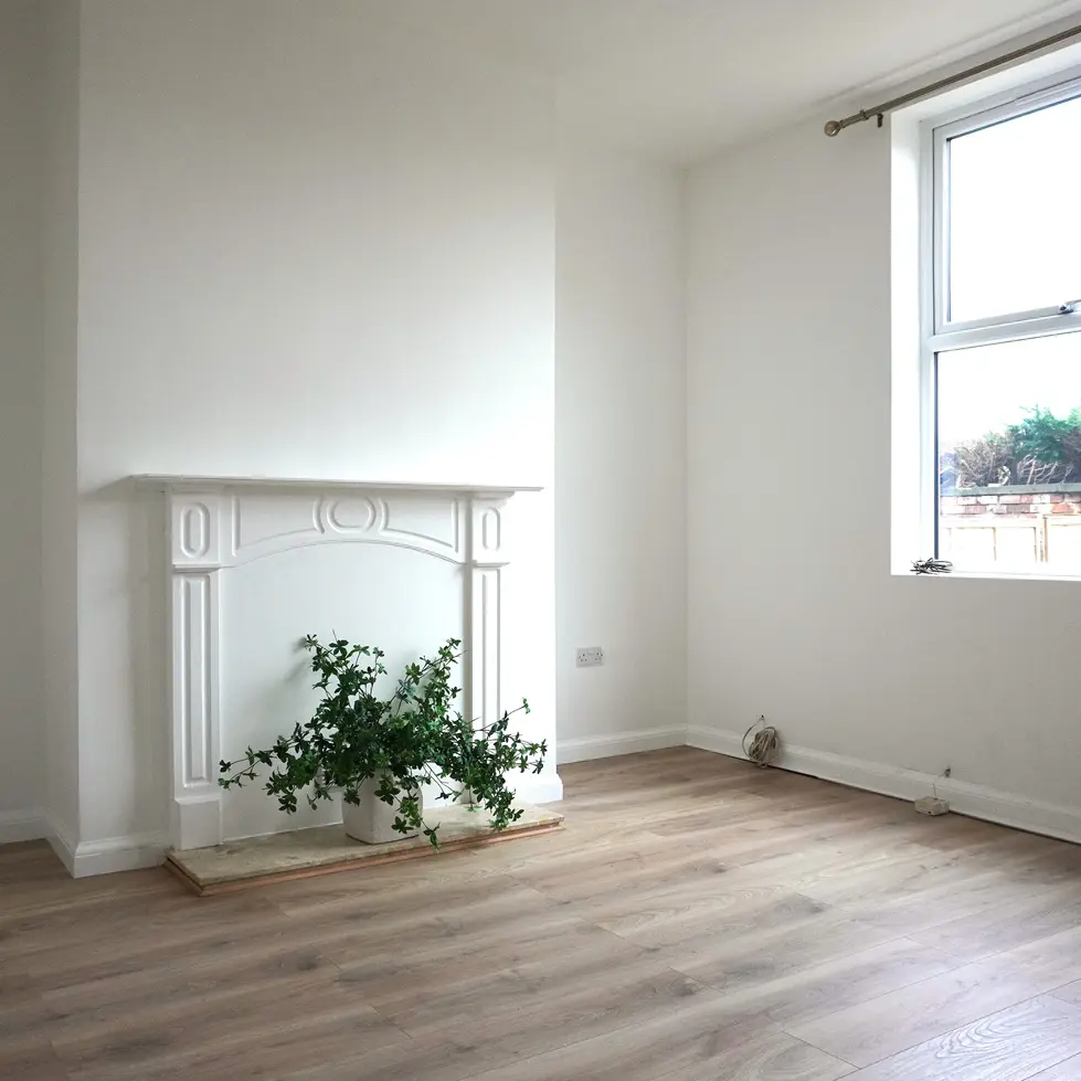 Refurbished living room with white walls, a decorative white fireplace, light wood flooring, and a potted plant, designed by Miriam Roberts.