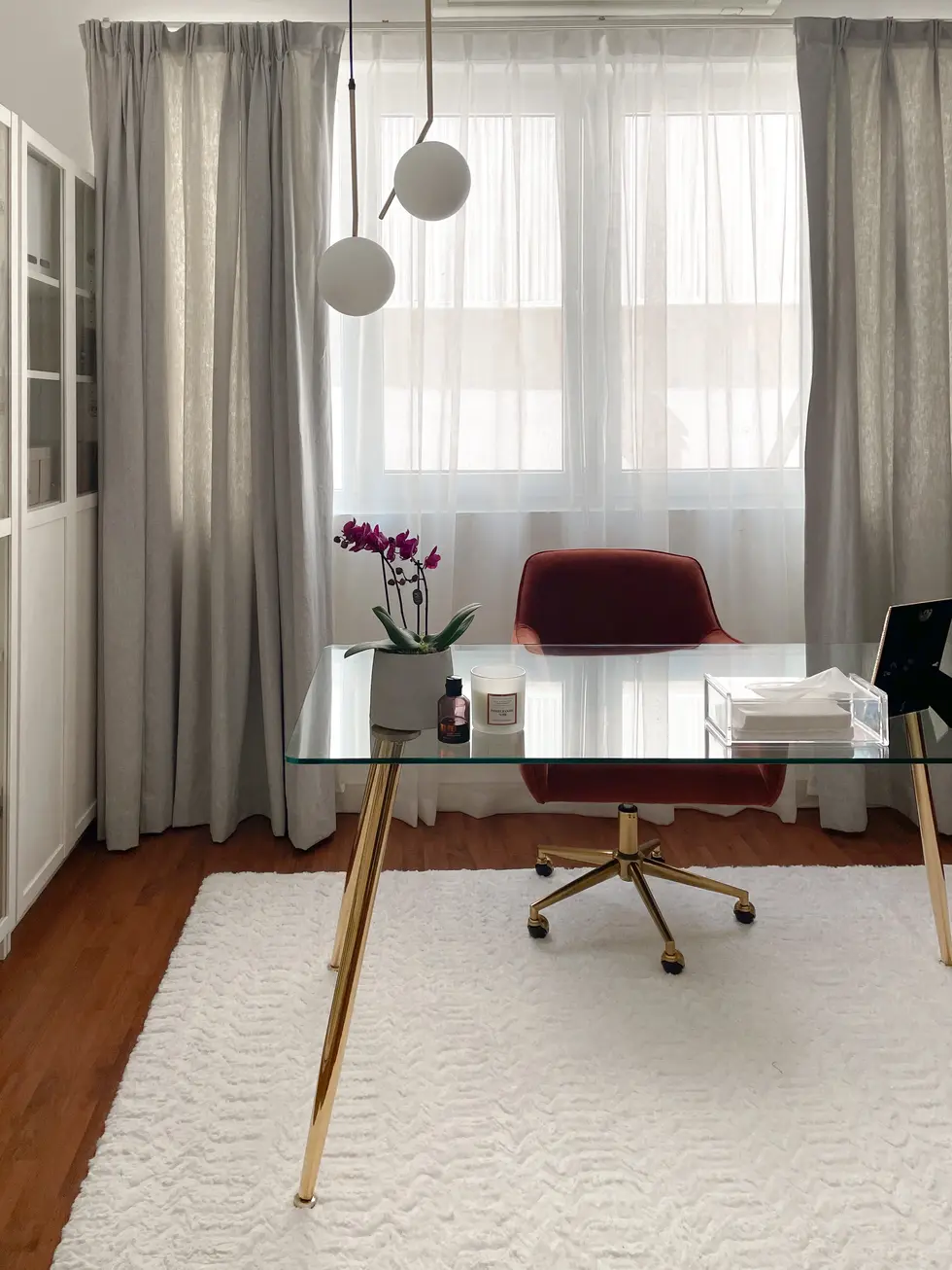 Sophisticated home office design by Miriam Roberts from the Roberts Renovations portfolio, featuring a glass-top desk with gold legs set on a plush white textured rug. A sleek red velvet chair with brass accents adds a touch of luxury, complemented by an orchid plant and minimal decor on the desk. Floor-to-ceiling grey curtains frame a large window, allowing soft natural light to fill the space. Modern pendant lights with globe shades hang above, enhancing the elegant and serene atmosphere. Rich wooden flooring contrasts beautifully with the light, airy tones of the room.