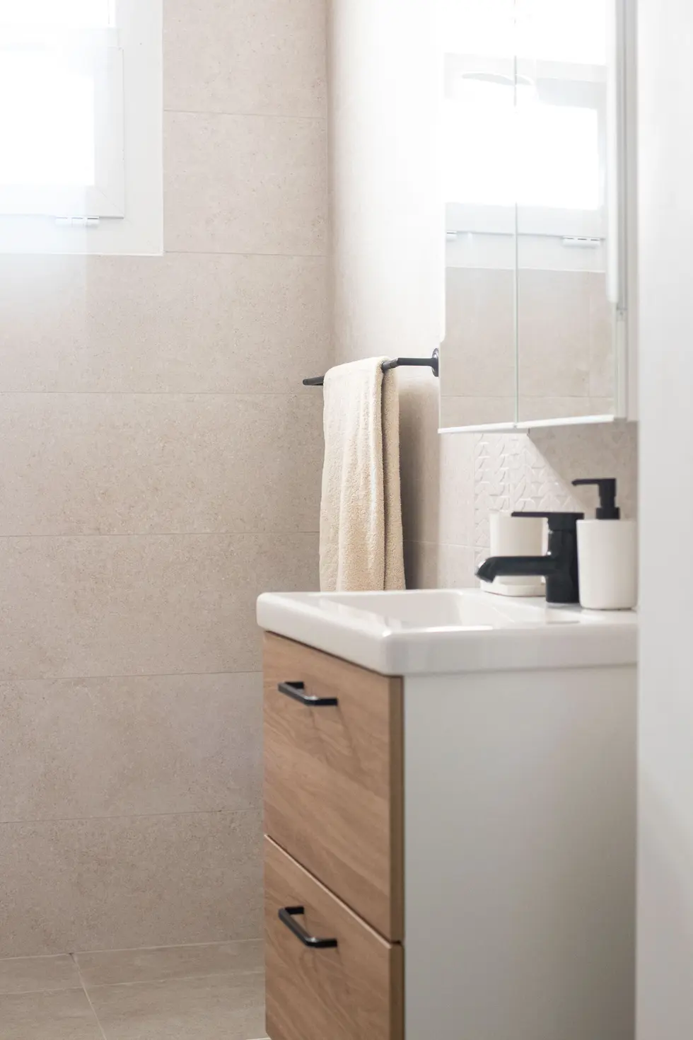 Minimalist bathroom with light beige tiles, wood vanity, black fixtures, and a mirrored cabinet, designed by Miriam Roberts for Roberts Renovation.