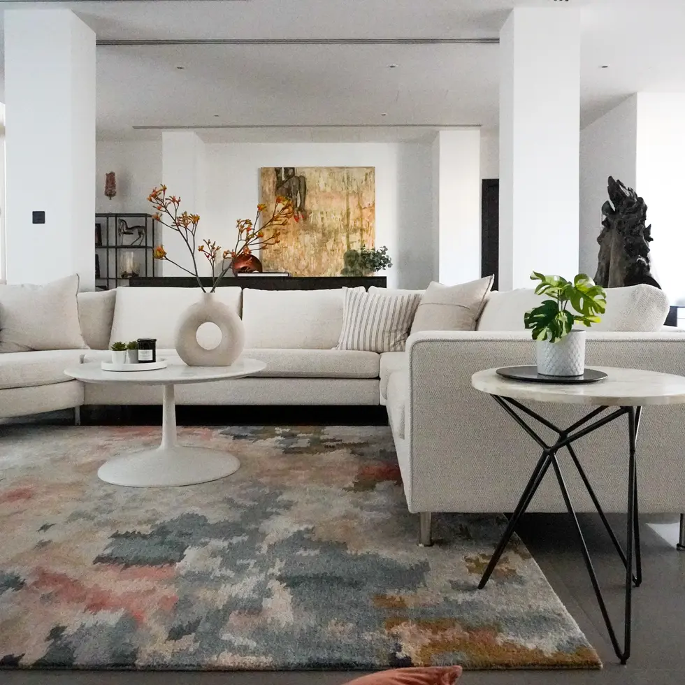 Contemporary living room design by Miriam Roberts from the Roberts Renovations portfolio, showcasing a spacious L-shaped beige sofa adorned with striped and plain cushions. The room features a round white coffee table with a minimalist vase holding orange-tinted branches and a scented candle, positioned over a multi-toned abstract rug. A side table with a white ceramic planter and green foliage complements the setting. In the background, an abstract artwork with warm tones hangs above a console, adding depth and character to the room. The open-plan layout and neutral palette create an airy, sophisticated atmosphere.