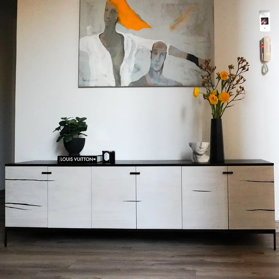 Modern living room design by Miriam Roberts from the Roberts Renovations portfolio, featuring a sleek black and white sideboard with minimalist handles and subtle wood grain patterns. The sideboard is adorned with a Louis Vuitton book, a black candle, and a potted plant for a touch of greenery. A tall black vase with vibrant yellow flowers adds a pop of colour alongside a sculptural white planter. Above the sideboard, a contemporary painting with abstract figures and warm orange accents draws attention, enhancing the room's artistic and refined atmosphere. The neutral walls and wooden flooring complete the elegant and curated look.