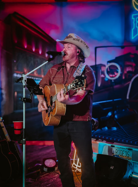 Ryan Evans wearing a UK Country hat at one of his gigs while playing a guitar and singing into a micraphone