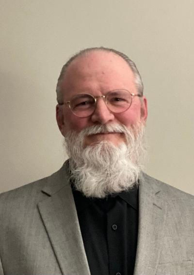 Portrait headshot of an older adult with glasses and a white beard in a light gray blazer and black shirt
