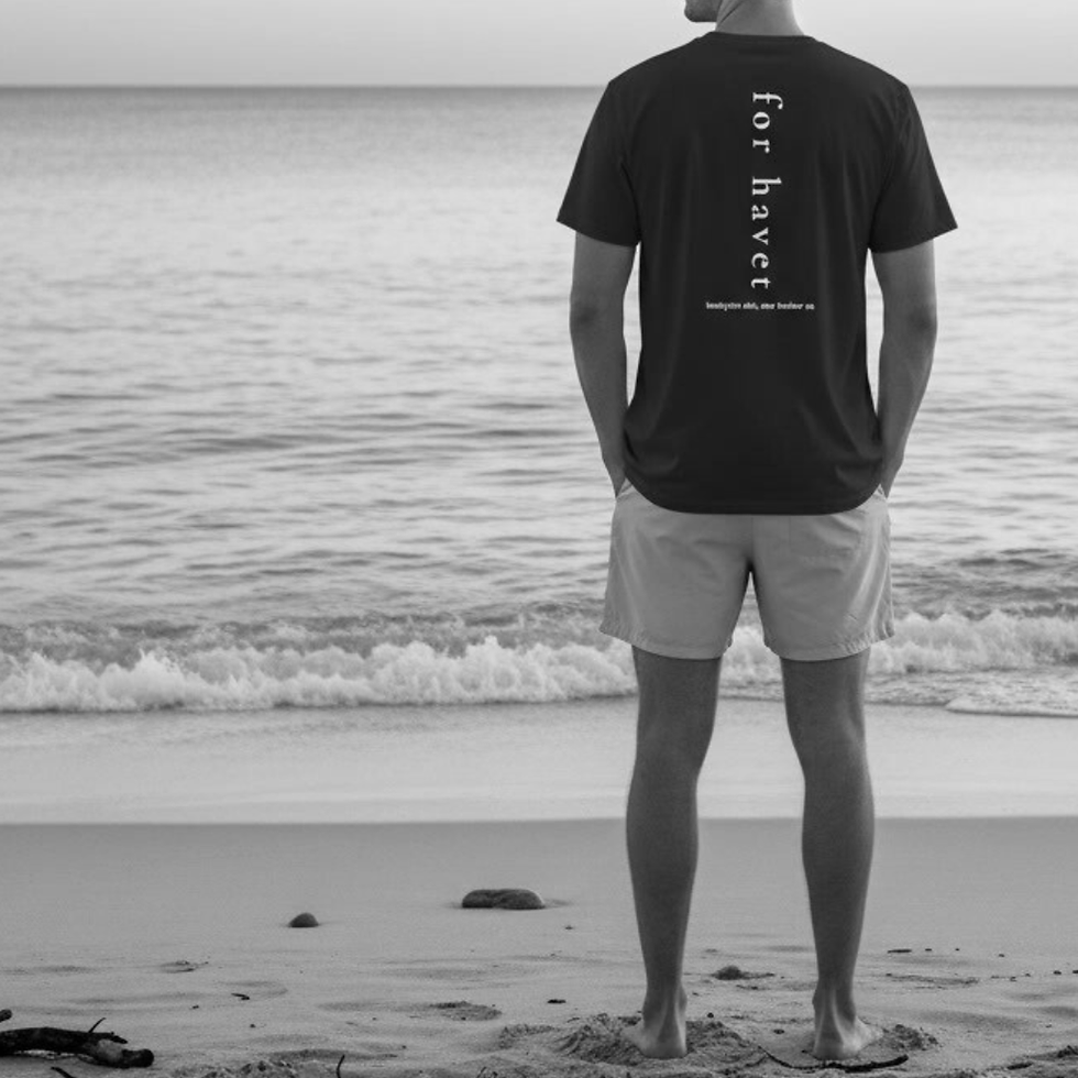 Man standing at the shoreline wearing a black William Bennett ‘For Havet’ tee. The back vertical text design is fully visible