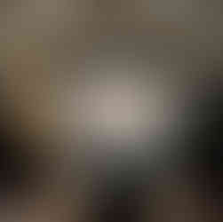 A dimly lit room with a monitor showing leather crafting. 3 teachers in apron are standing in front, one of them is speaking into a mic. there are some people sitting at a round table watching the monitor.