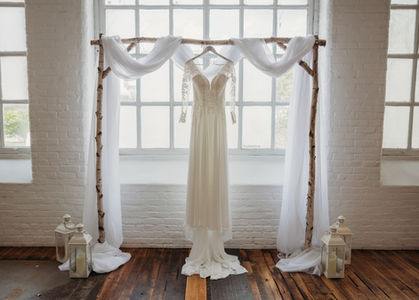 Birch Arch with draping across the top and white lanterns on the floor. Photo shows the wedding dress hanging on the arch.