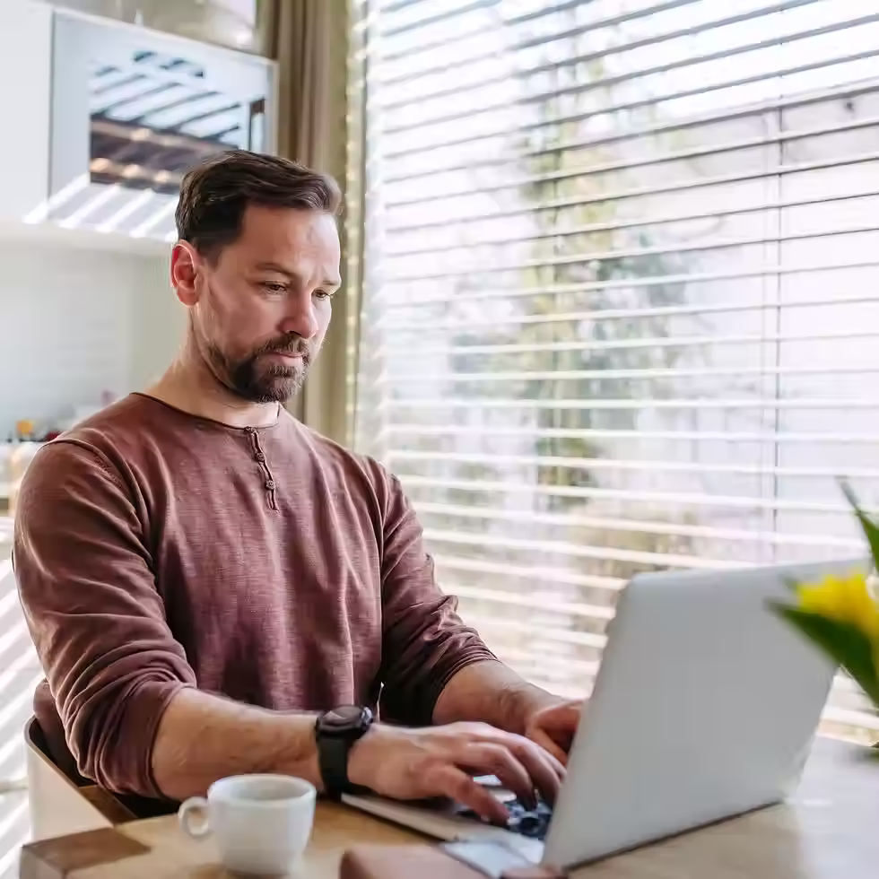 A man works on a laptop at a home desk beside a window with blinds.