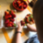 A bowl of cut strawberries sits at the top of the image while the pack of whole strawberries sits to the left. A boy's hands can be seen cutting a strawberry with an green-handled knife.