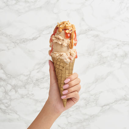 A hand with neatly manicured nails holds a waffle cone stacked with scoops of ice cream, topped with drizzled peanut butter and crushed peanuts against a marble background.