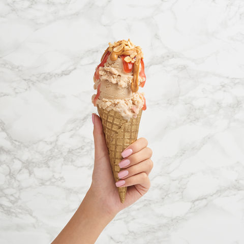 A hand with neatly manicured nails holds a waffle cone stacked with scoops of ice cream, topped with drizzled peanut butter and crushed peanuts against a marble background.