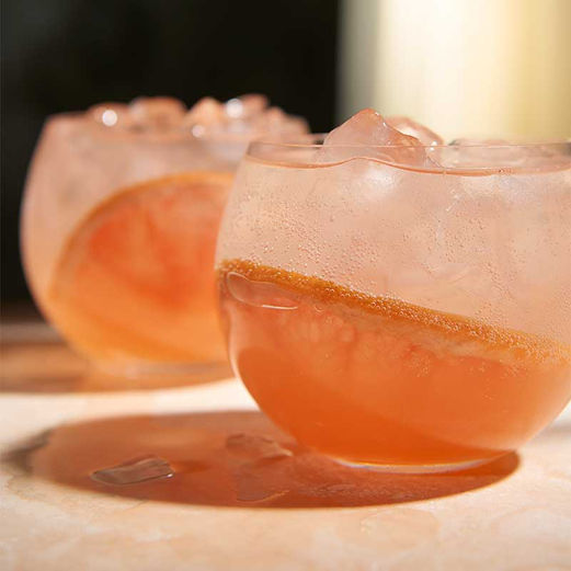 Two round glasses of peach-colored cocktails with large ice cubes
