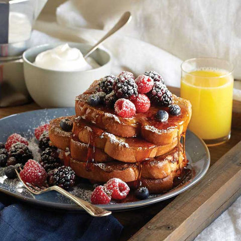 A stack of golden French toast drizzled with syrup and topped with fresh raspberries, blackberries, and blueberries, dusted with powdered sugar. A side of whipped cream, orange juice, and a fork rest nearby on a breakfast tray.