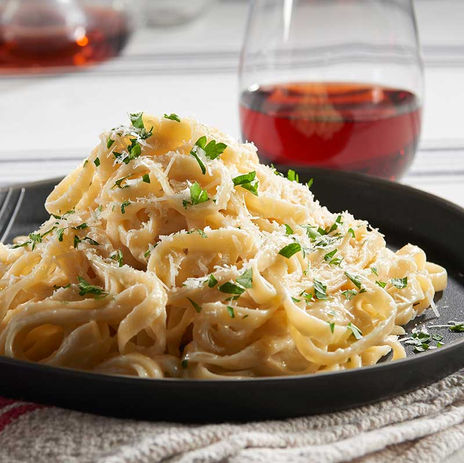A plate of creamy fettuccine Alfredo topped with freshly grated Parmesan and chopped parsley, served on a black dish with a glass of red wine in the background.