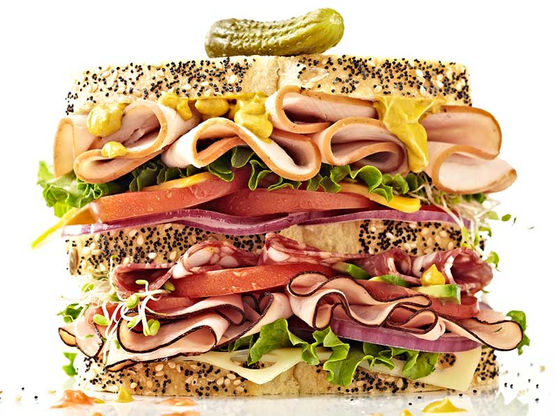 A towering deli sandwich made with poppy seed bread, layered with thinly sliced turkey and ham, crisp lettuce, tomatoes, red onions, sprouts, Swiss cheese, and mustard, topped with a pickle.