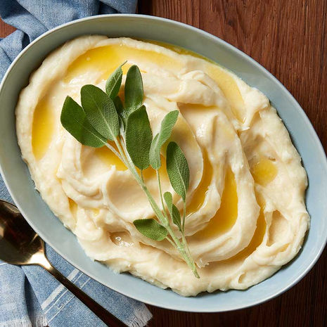 A bowl of smooth mashed potatoes swirled with melted butter and garnished with fresh sage, set on a wooden table with a blue napkin and gold spoon.