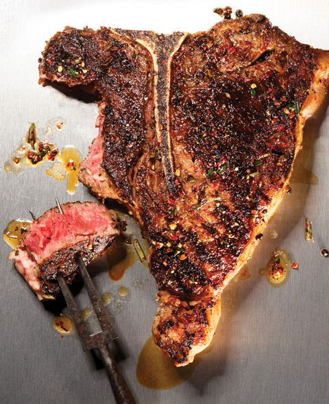A juicy, spice-crusted T-bone steak with a charred exterior sits on a metal surface, with a tender, medium-rare slice cut and held by a meat fork. Drizzles of rendered fat and seasoning flakes surround the steak.