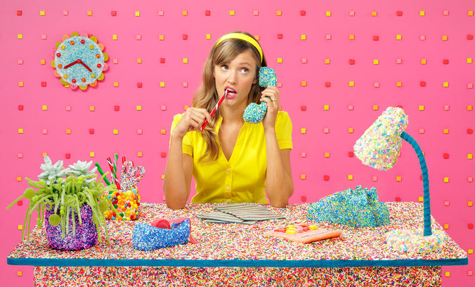 A woman in a yellow outfit sits at a desk covered in colorful sprinkles, holding a blue sprinkle-covered phone and biting a red pencil. The background features a pink wall with a sprinkle-covered clock and various candy-coated office supplies.