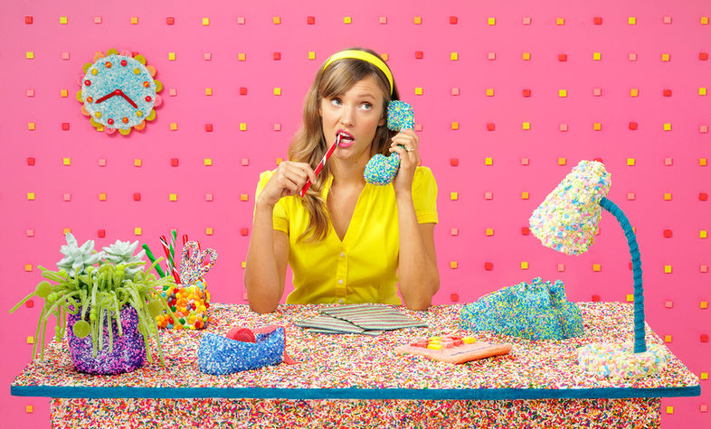 A woman in a yellow outfit sits at a desk covered in colorful sprinkles, holding a blue sprinkle-covered phone and biting a red pencil. The background features a pink wall with a sprinkle-covered clock and various candy-coated office supplies.