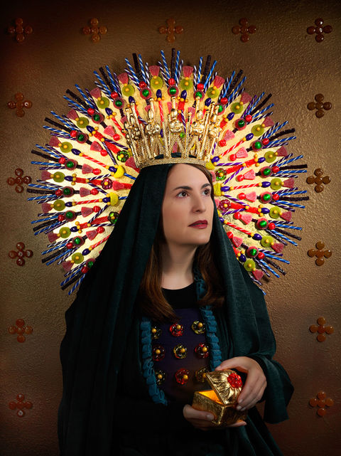 A woman dressed in dark, regal robes wears an ornate halo-like crown made of colorful lollipops and candy sticks, holding a gilded gift against a rich, textured background.