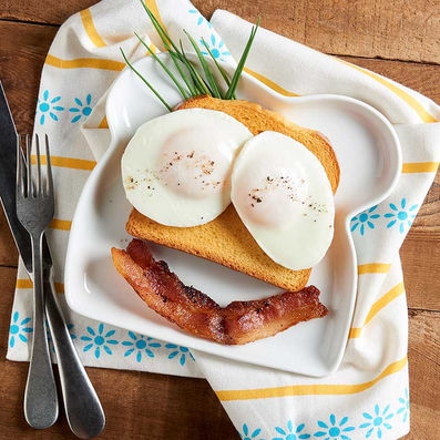 A playful breakfast arrangement featuring two sunny-side-up eggs on toast as eyes, a strip of crispy bacon as a smile, and chives as eyebrows, served on a toast-shaped plate with a patterned napkin.